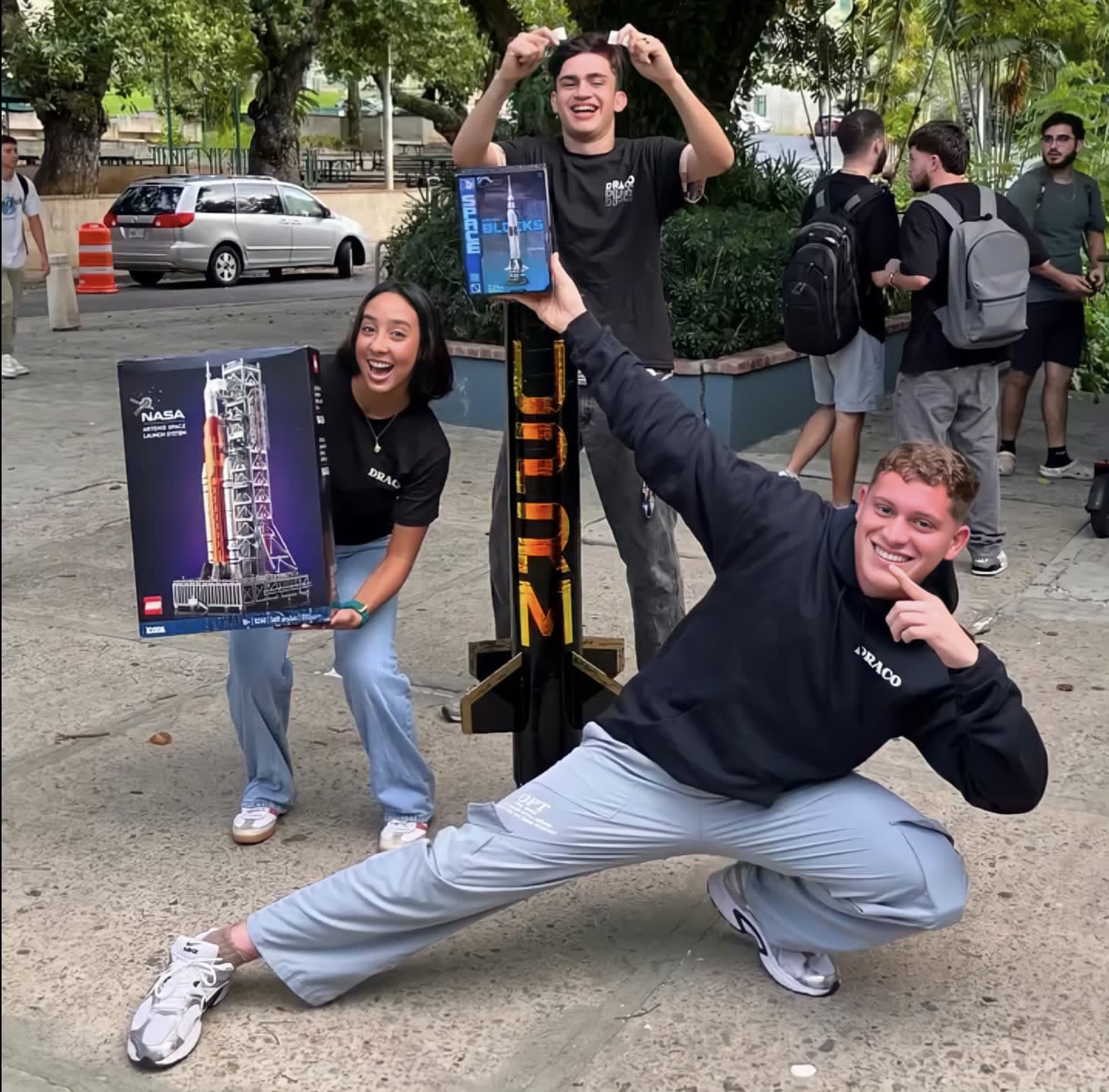 Three DRACO members posing on the UPRM campus holding LEGO NASA rocket sets beside a rocket sculpture