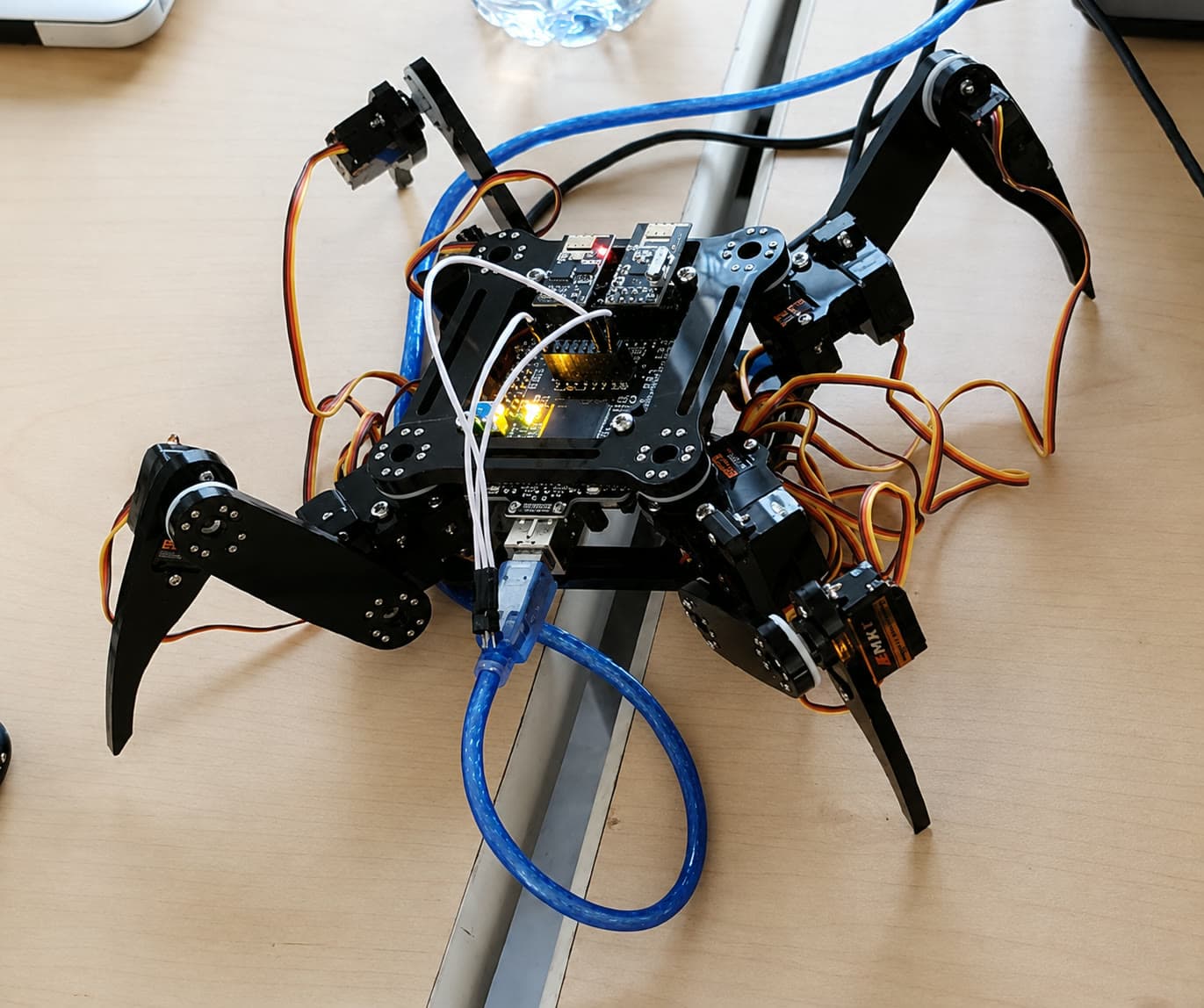 Six-legged robot with black articulated legs, servo motors, and a microcontroller board wired on a workbench