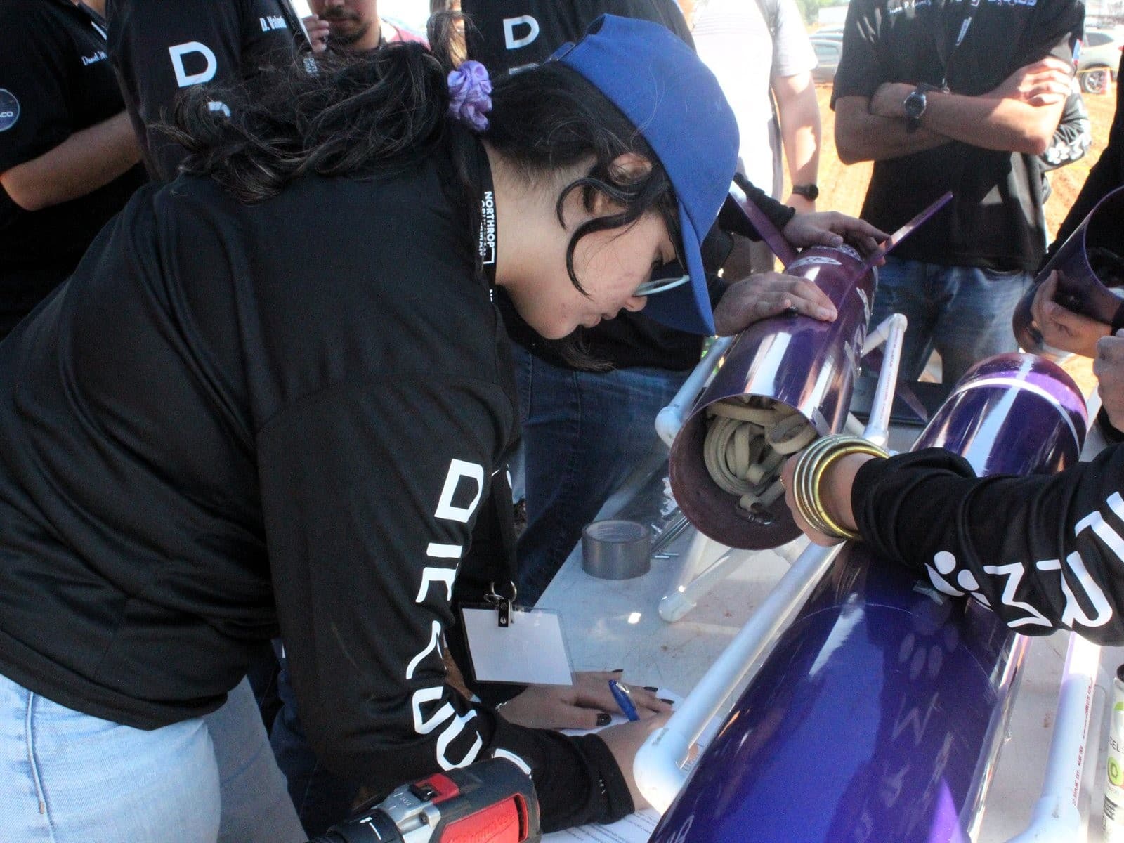 A DRACO member filling out a written assembly checklist at the launch field while the team works on the purple rocket airframe beside her