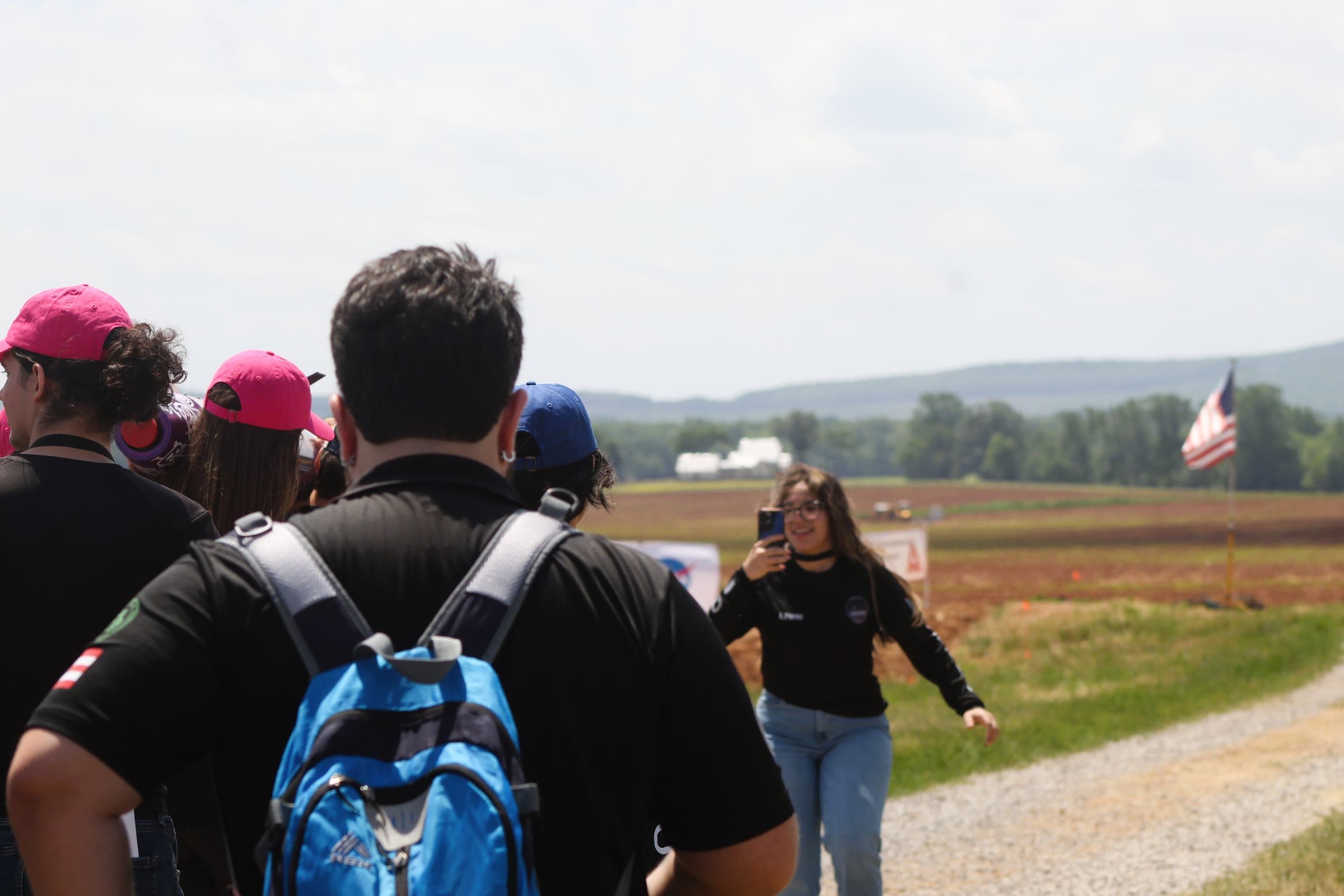 DRACO members walking along a dirt road at the launch field while a teammate films them, with an American flag and the range in the background