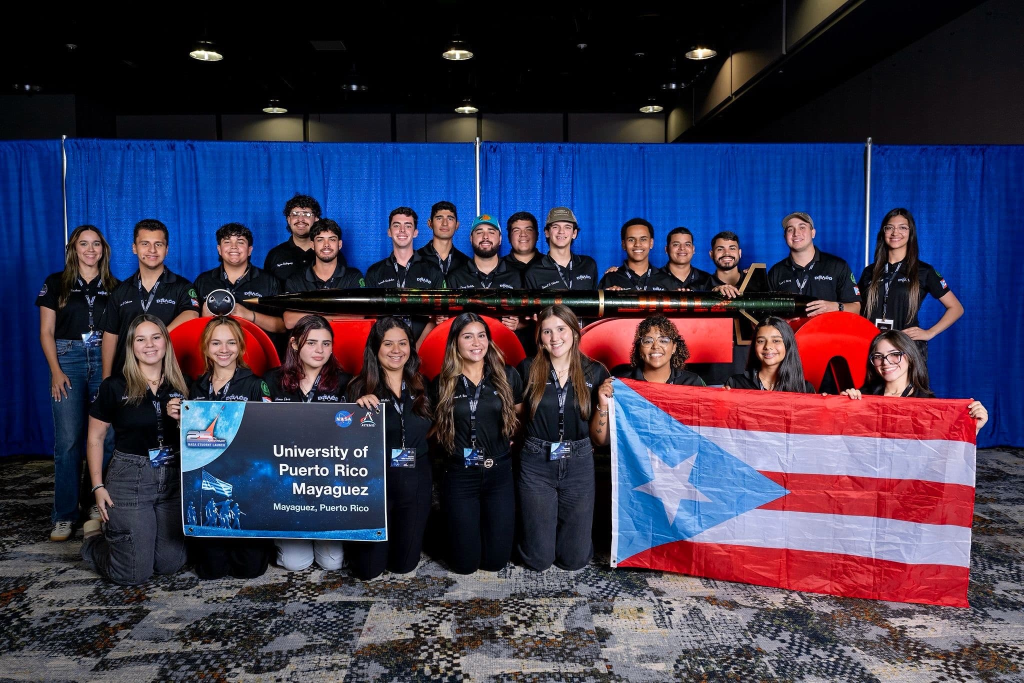 The 2025 DRACO team at the competition venue holding the Forest Wyvern airframe, a University of Puerto Rico at Mayagüez sign, and a Puerto Rico flag